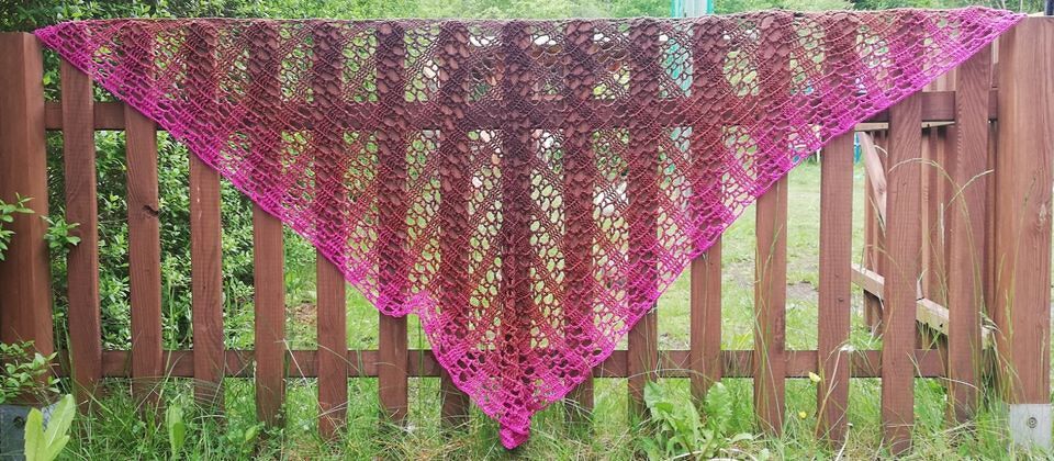 Gehäkeltes Dreieckstuch in Pink‑ und Brauntönen mit offenem Lochmuster, aufgehängt an einem Holzzaun