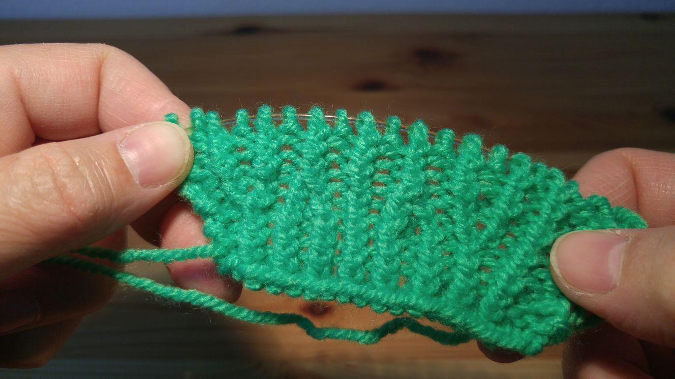 Hände halten grünes Strickstück mit reliefartigem Strukturmuster auf Rundstricknadel