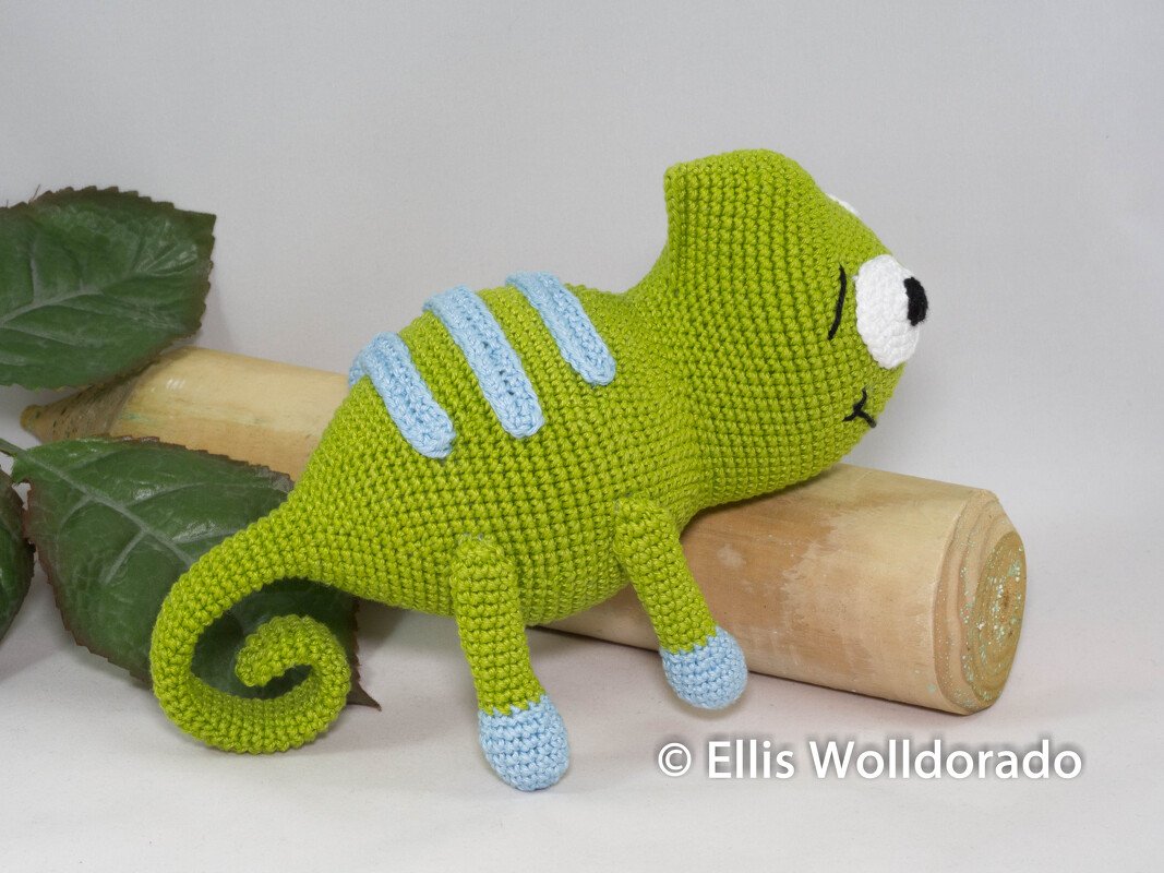 Gehäkeltes grünes Chamäleon‑Amigurumi mit hellblauen Streifen und eingerolltem Schwanz auf Holz