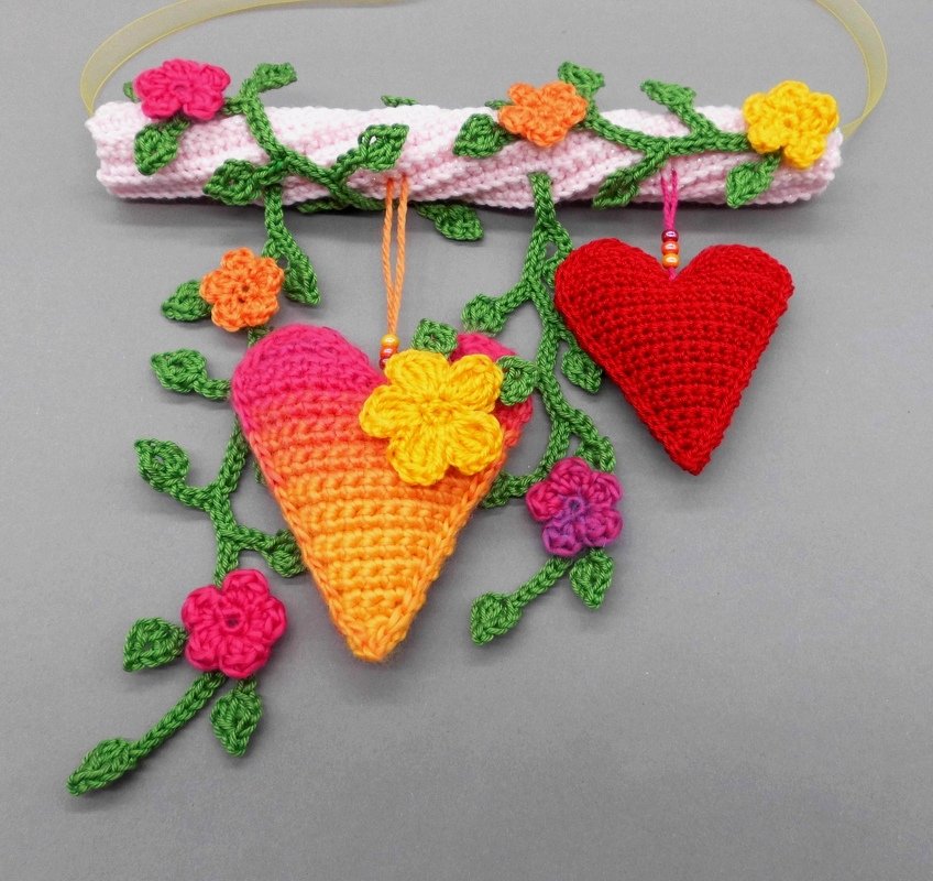 Crocheted wall hanging with red and pink-orange hearts, green vines, and small colorful flowers on a pink bar.