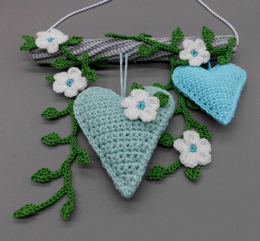 Two light-aqua crocheted hearts hanging from a gray crocheted rod with green crochet vines and white crochet flowers