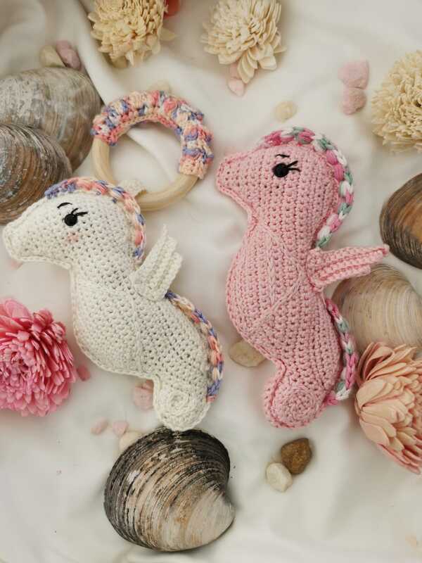 Zwei gehäkelte Seepferdchen (weiß und rosa) und ein Greifring aus Holz mit Häkelbezug auf hellem Stoff, umgeben von Muscheln und Trockenblumen.