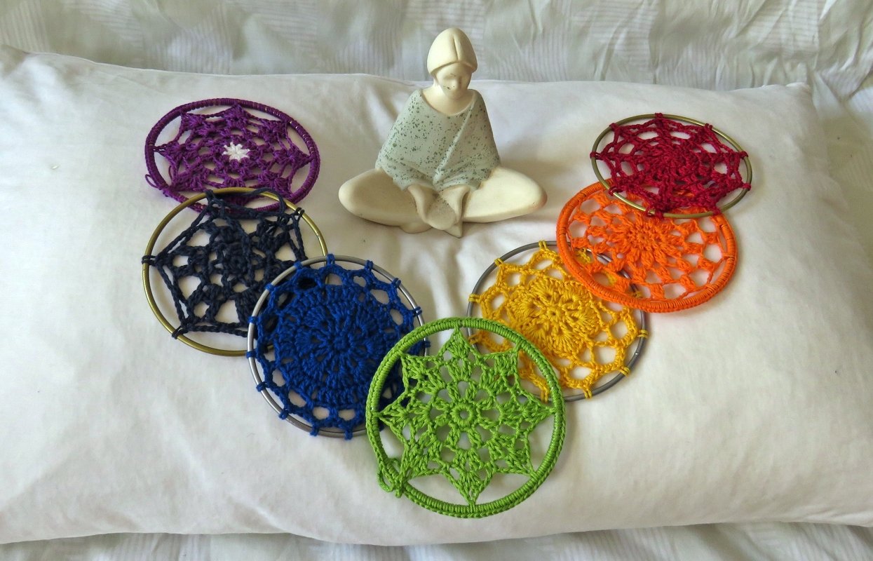 Halbkreis aus gehäkelten Kreis‑Motiven in Metallringen in Regenbogenfarben auf heller Stoffunterlage neben einer sitzenden Keramikfigur