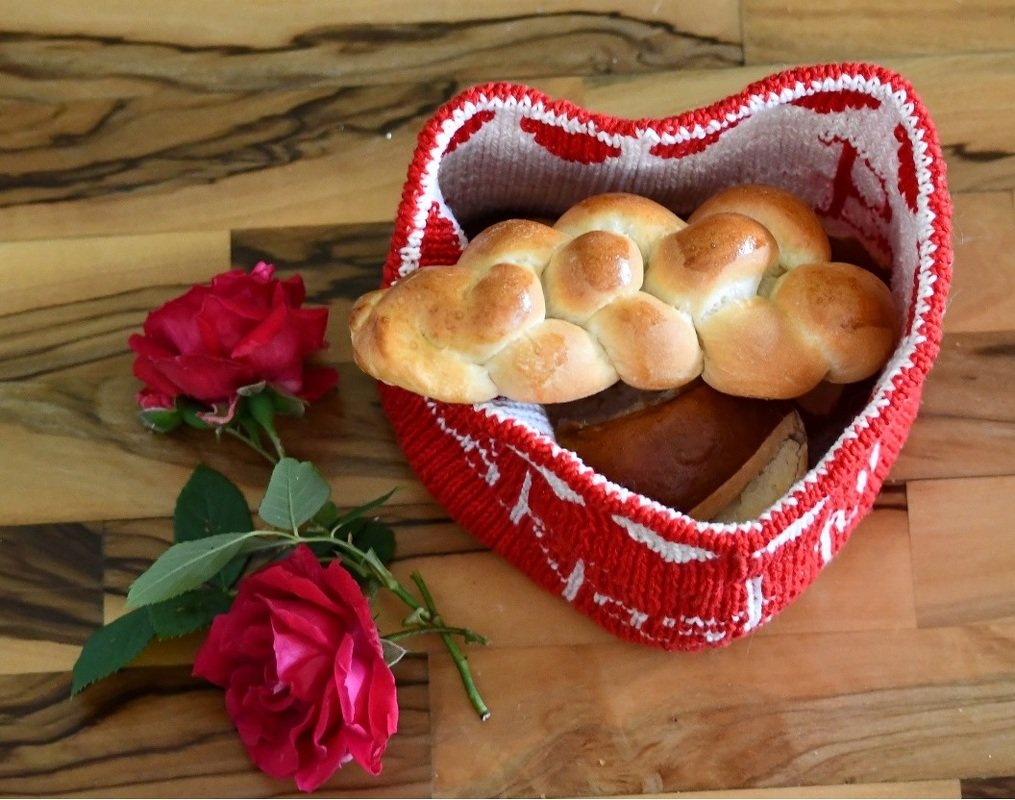 Gestrickter Herzkorb in Rot und Weiß mit Zopfbrot und zwei roten Rosen auf Holzhintergrund.