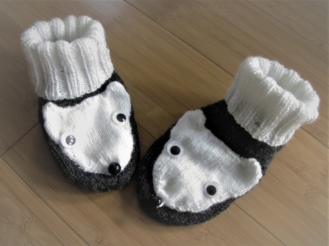 Paar schwarz-weißer, gestrickter Babyschuhe mit Tiergesicht und Wackelaugen auf hellem Holzfußboden.