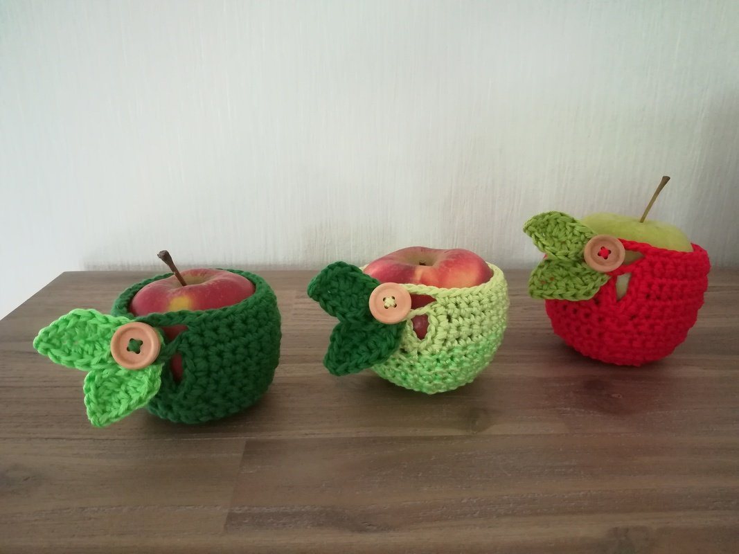 Drei gehäkelte Apfel-Utensilos in Grün und Rot mit gehäkelten Blättern und Holzknöpfen, gefüllt mit Äpfeln auf einem Holztisch.