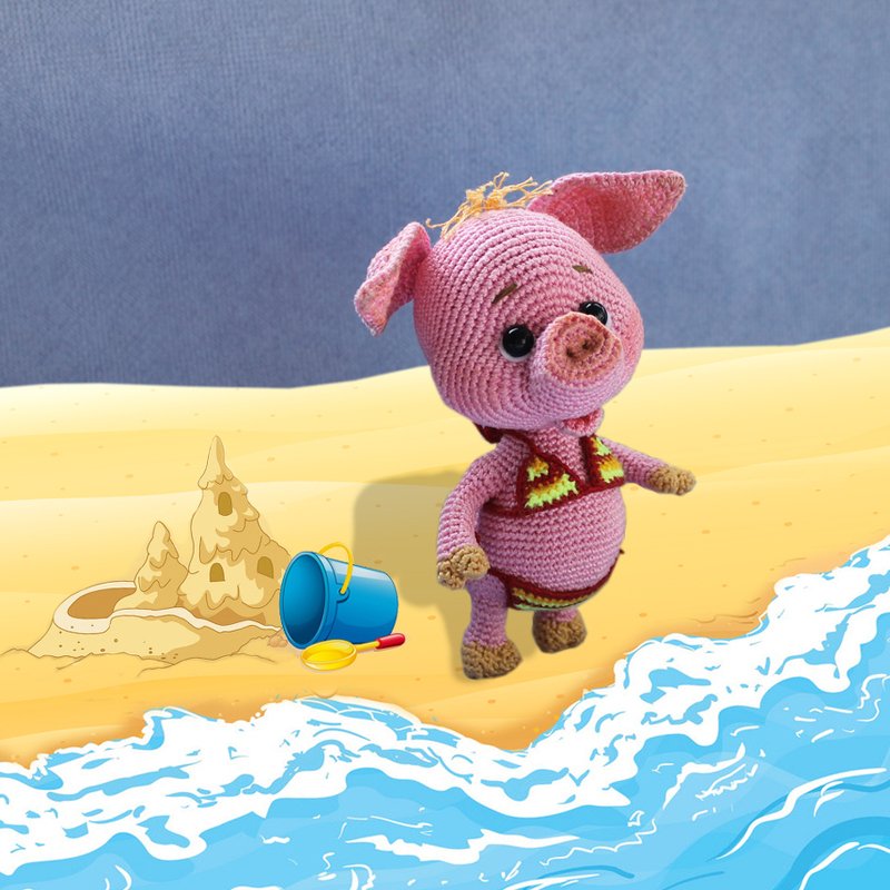 Gehäkeltes rosa Amigurumi-Schwein mit rotem Bikini am Strand neben Sandburg, Eimer und Wellen