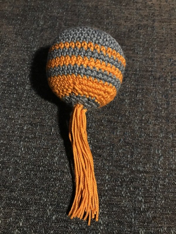 Gehäkelter grauer und oranger Katzenball mit langer Quaste auf dunklem Stoff