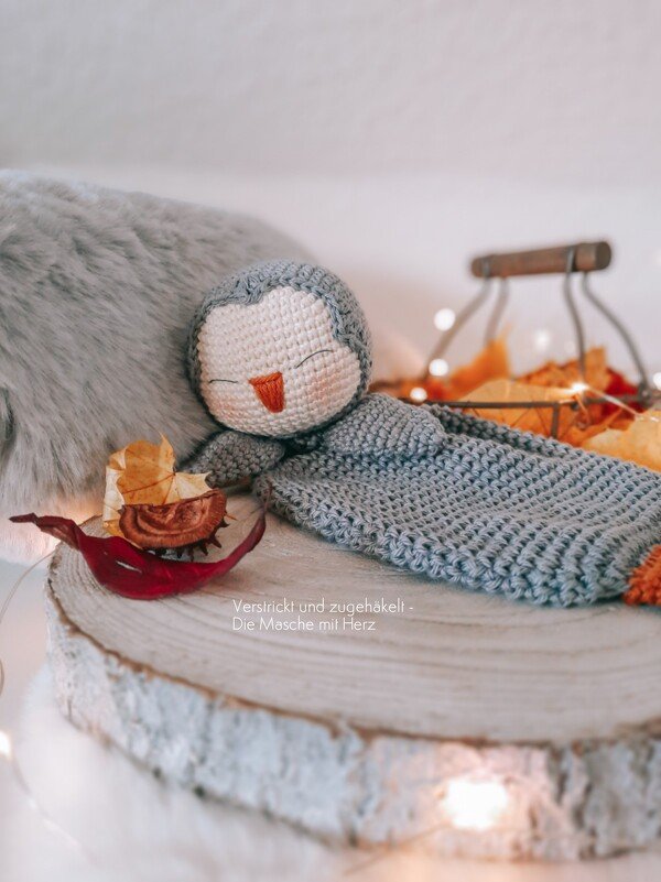 Gray and white crocheted penguin baby comforter on a wooden slice with autumn leaves and fairy lights.