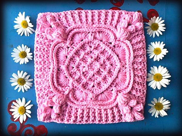 Rosa gehäkeltes Quadrat mit mittiger Blüte, Noppen- und Zopfmuster, umgeben von Gänseblümchen auf blauem Hintergrund.