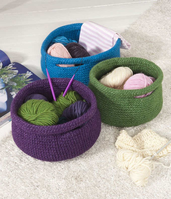 Drei gehäkelte Körbe in Lila, Blau und Grün mit Wollknäueln und Stricknadeln auf hellem Teppich.