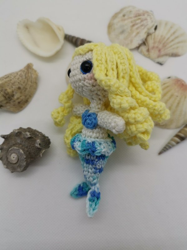 kleine gehäkelte Meerjungfrau mit gelbem Lockenhaar und blau-türkisfarbener Flosse neben Muscheln