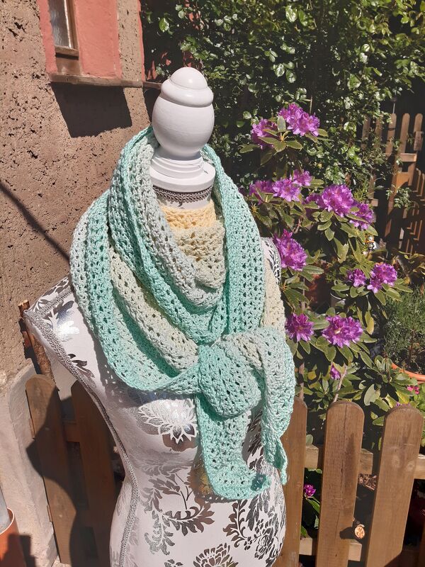 Häkel-Tuch in Pastellfarben auf einer Puppe drapiert im Garten neben lila Blüten