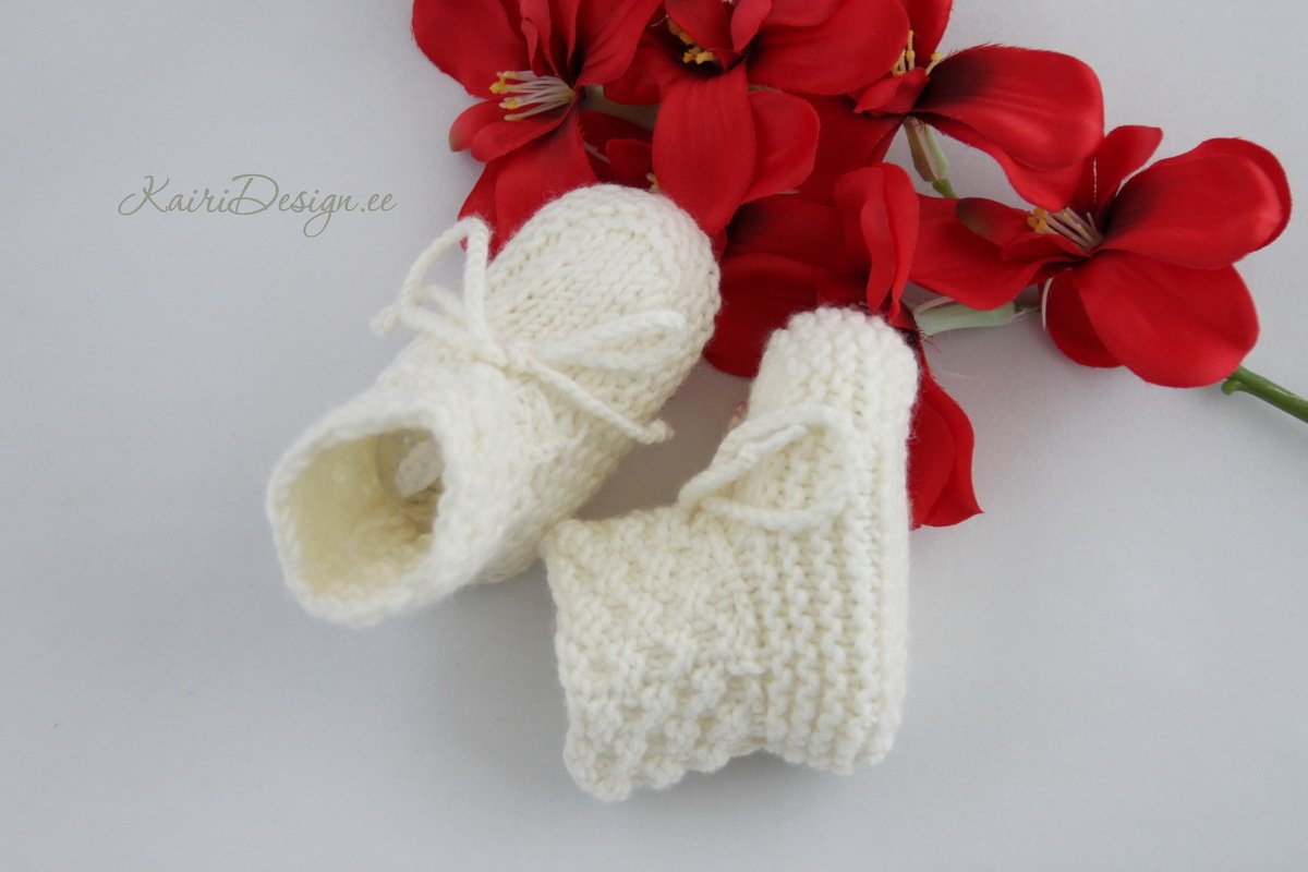 Cream hand-knit baby booties with tie cords beside red flowers