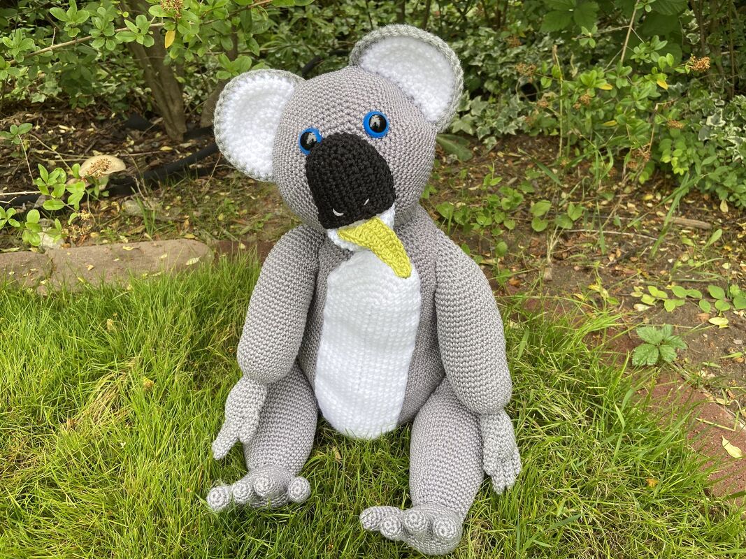 Gehäkelter grauer Koala mit weißem Bauch, schwarzer Nase und blauen Augen, sitzt auf Gras und hält ein gelb-grünes Blatt im Mund.