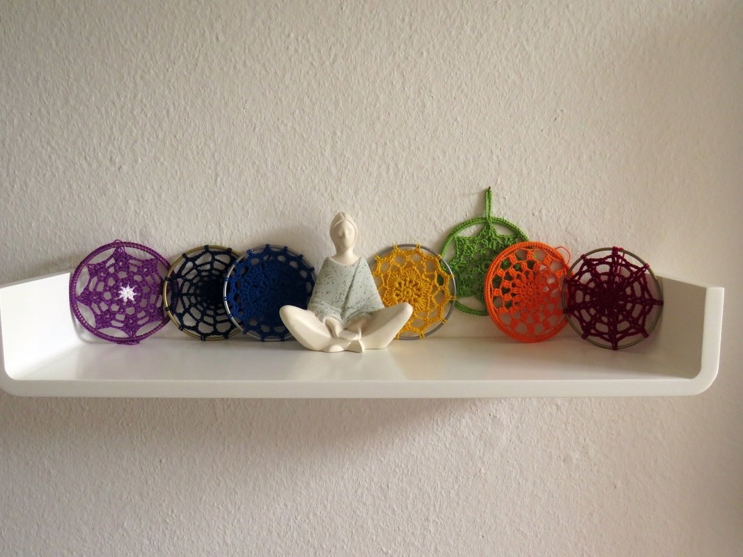 Weiße sitzende Meditationsfigur umgeben von bunten, gehäkelten Mandalas auf einem weißen Regal.