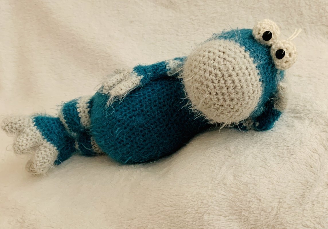 Blau-creme gehäkeltes Amigurumi-Fantasiewesen liegend mit großer runder Schnauze und Kulleraugen