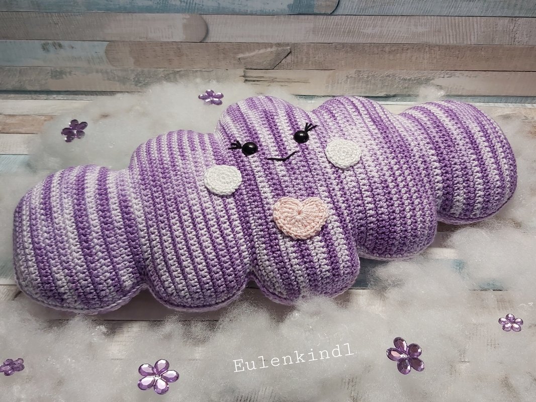 Gehäkeltes Wolkenkissen, lila-weiß gestreift, mit aufgesticktem Gesicht und rosa Herz, auf weicher Unterlage liegend.