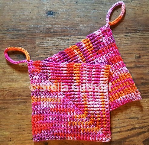 Zwei gehäkelte Topflappen in Pink- und Orangetönen mit Schlaufen auf Holzuntergrund