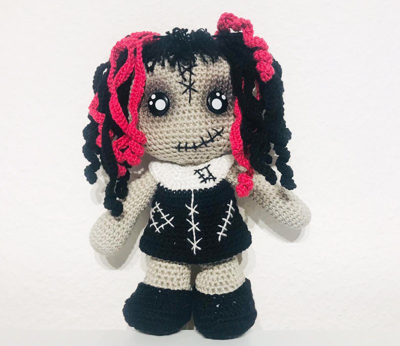 Gehäkelte Amigurumi-Puppe mit grauer Haut, schwarzem Kleid, weißen aufgestickten Nähten und pink-schwarzen lockigen Haaren