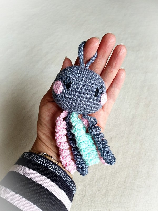 Gehäkelte graue Amigurumi-Qualle mit rosa Bäckchen und bunten spiralförmigen Tentakeln, gehalten in einer Hand.