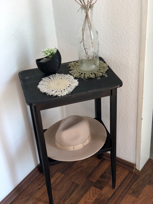 Zwei gehäkelte Untersetzer (hellbeige und olivgrün) auf dunklem Beistelltisch mit Glasvase, getrockneten Zweigen und kleinem Blumentopf.