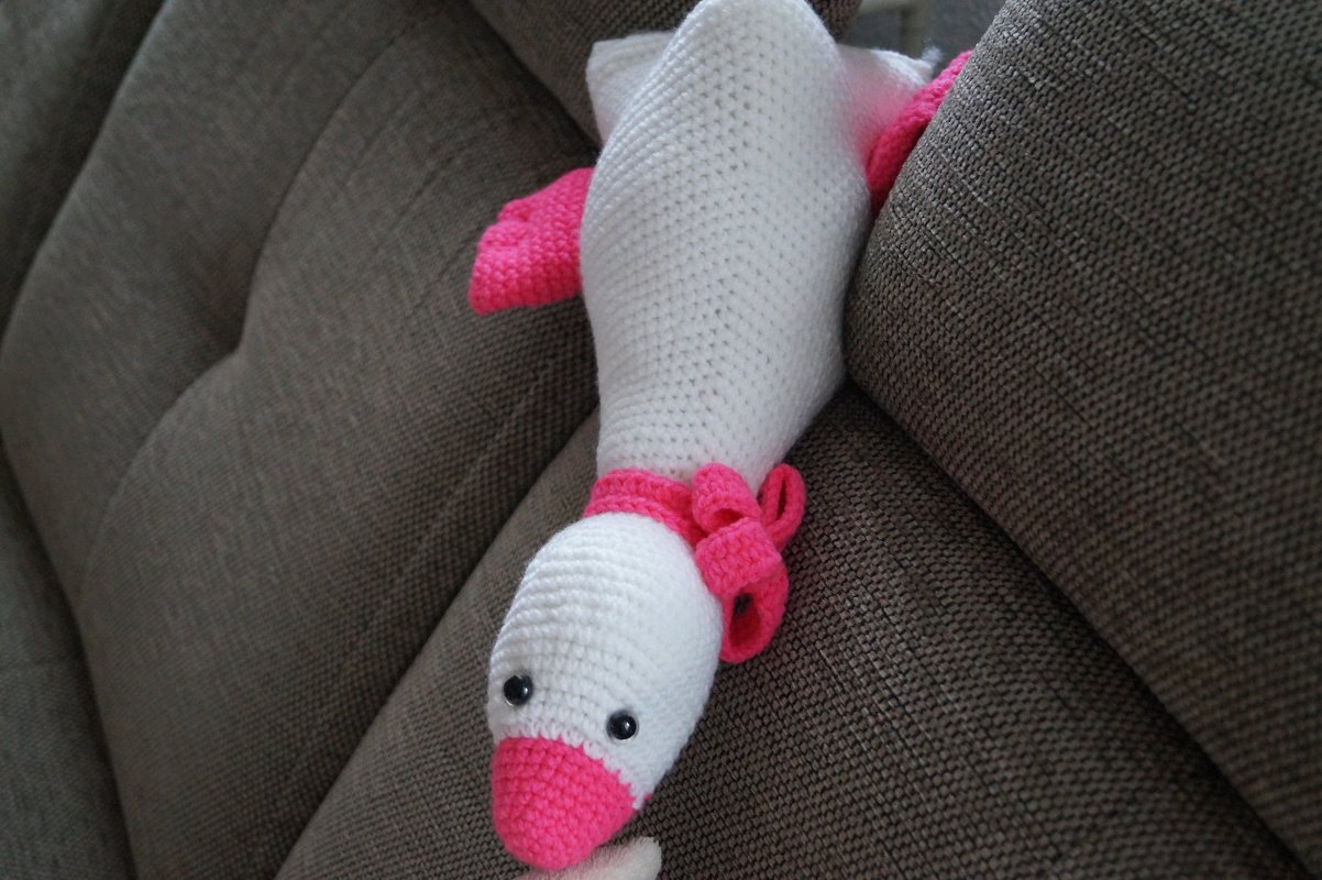 Nahaufnahme einer weißen gehäkelten Amigurumi-Gans mit pinkem Schnabel, pinken Flügeln und kleinen schwarzen Augen auf braunem Sofa
