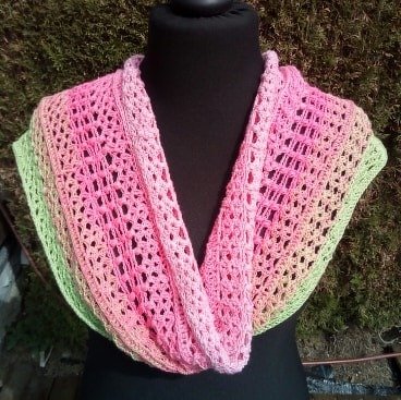Gehäkelter Loop‑Schal in Pink und Grün mit offenem Muster auf schwarzer Schneiderpuppe vor grüner Hecke