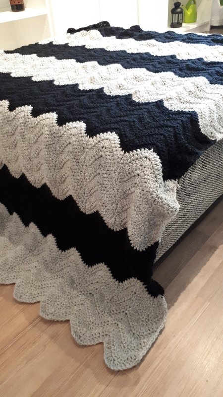 Gehäkelte Decke mit Zickzackmuster in Hellgrau und Dunkelblau, über die Bettkante gelegt.