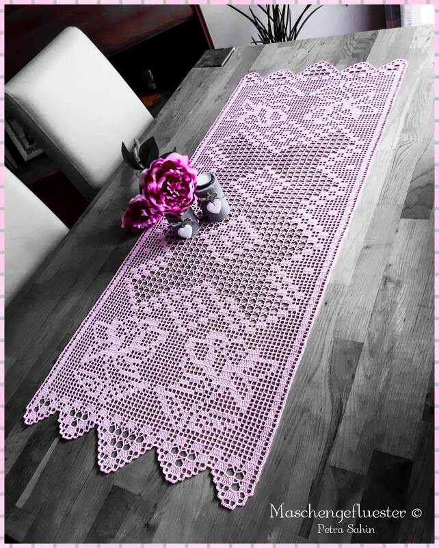 Rosa gehäkelter Filet-Tischläufer mit Blumenmuster und gezacktem Rand auf Holztisch, dekoriert mit Blüten