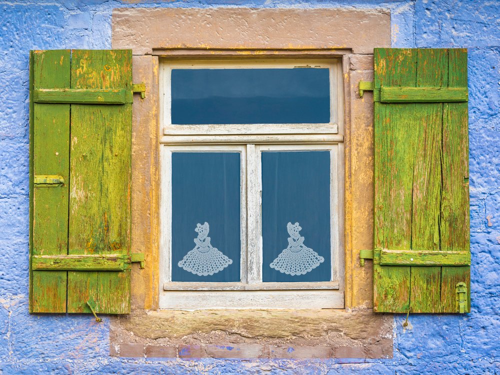 Fenster mit verwitterten gr
Rnen Fensterläden, blauer Putz und zwei zarten weißen Häkelpuppen als Dekor an der Scheibe