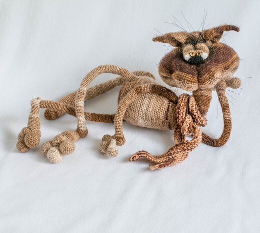 Gehäkelte Amigurumi-Katze in Braun- und Beigetönen, liegend auf hellem Hintergrund, mit großem Kopf und langen Beinen.