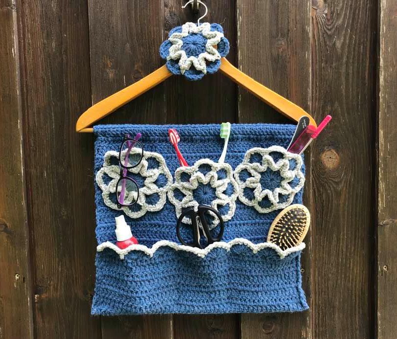 Blauer gehäkelter Wand-Organizer mit Blumenapplikationen und Taschen, aufgehängt an einem Holzkleiderbügel, gefüllt mit Alltagsgegenständen.