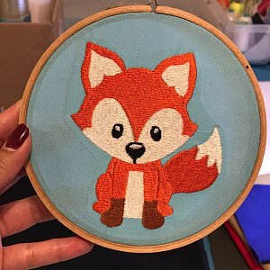 Roter cartoonhafter Fuchs mit weißer Brust und Schwanzspitze in einem Holzstickrahmen auf hellblauem Stoff