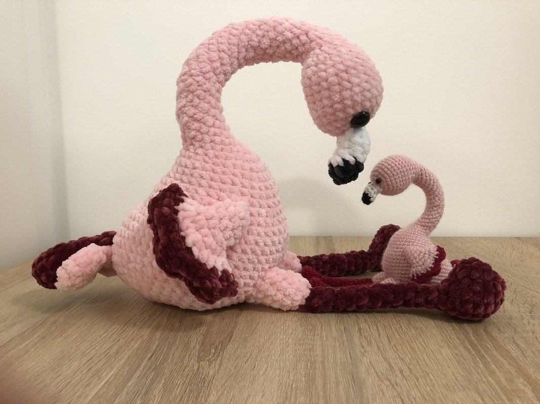 Zwei gehäkelte Flamingos, groß und klein, aus rosa Flauschgarn mit dunkelroten Akzenten auf einem Holztisch