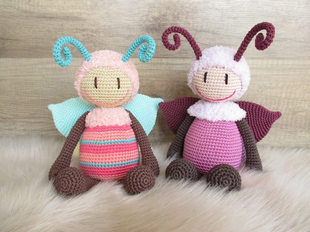 Zwei Amigurumi-Figuren mit verschiedenen Garntexturen, Flügeln und spiralförmigen Fühlern in Pastellfarben