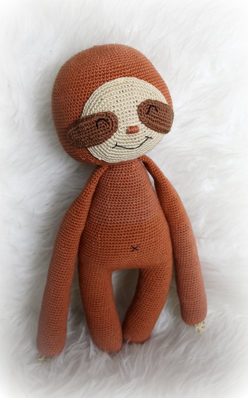 Gehäkeltes braunes Faultier-Amigurumi mit heller Gesichtsmaske, langen Armen und lächelnder Bestickung auf weißem Plüsch