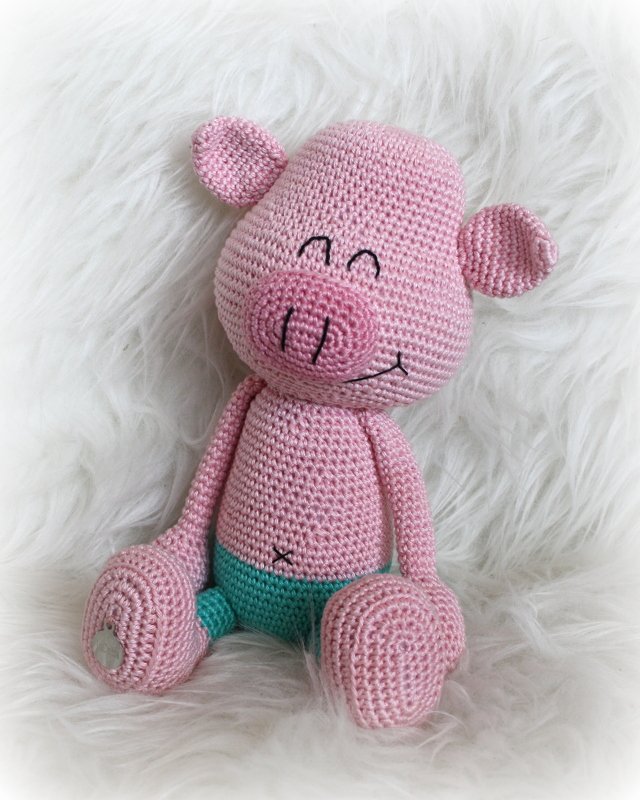 Rosa gehäkeltes Amigurumi‑Schwein mit türkisfarbener Hose auf weißem Fell