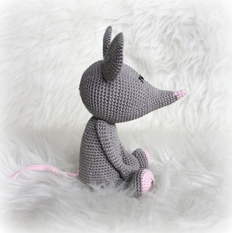 Gehäkelte graue Amigurumi‑Maus mit rosa Nase, Ohren und Pfoten auf weißem, flauschigem Untergrund, seitlich gesehen.