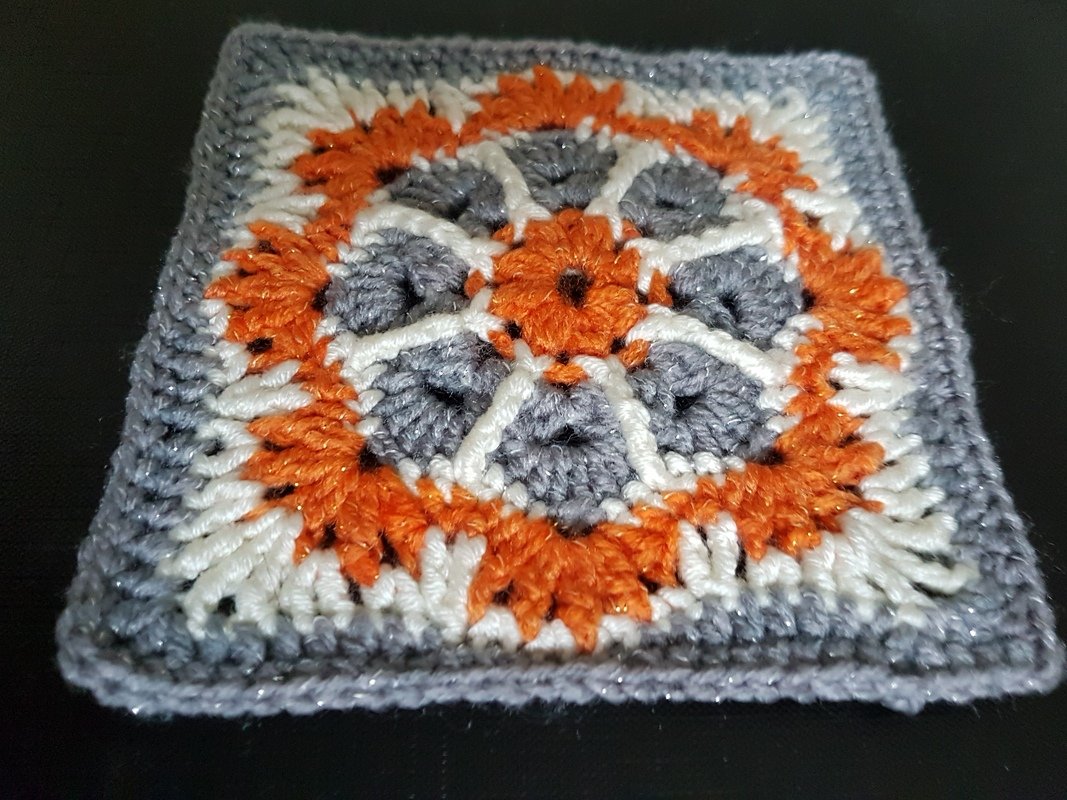 Quadratisches Häkelmotiv mit orange-grau-weißem, glitzerndem Blumenmuster und strukturierter Umrandung