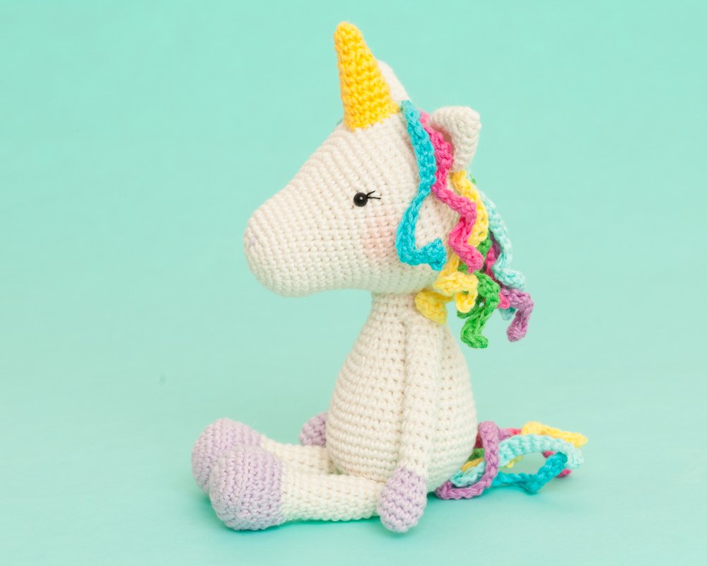 Cream crocheted amigurumi unicorn with yellow horn and pastel curly mane sitting on a mint background