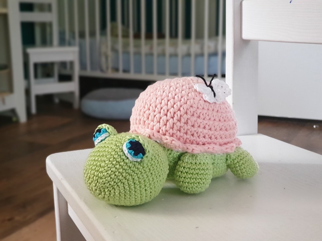 Gehäkelte hellgrüne Schildkröte mit rosa Panzer und weißem Schmetterling, sitzend auf weißem Stuhl