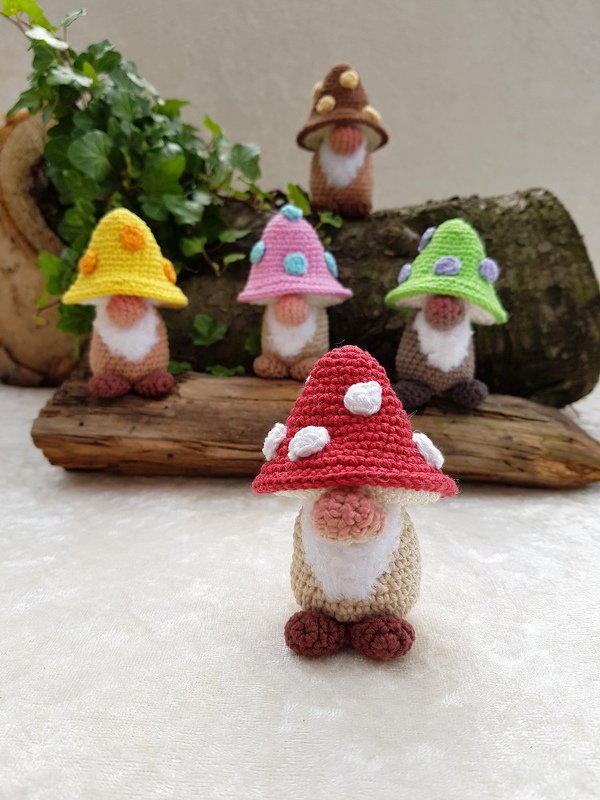 Nahaufnahme eines roten gehäkelten Pilz‑Wichtels im Vordergrund; mehrere bunte Wichtel mit Pilzhüten im Hintergrund.