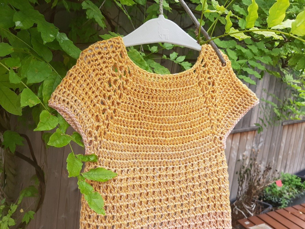 Gehäkeltes gelbes Kinderkleid mit Lochmuster auf weißem Bügel vor grünen Blättern