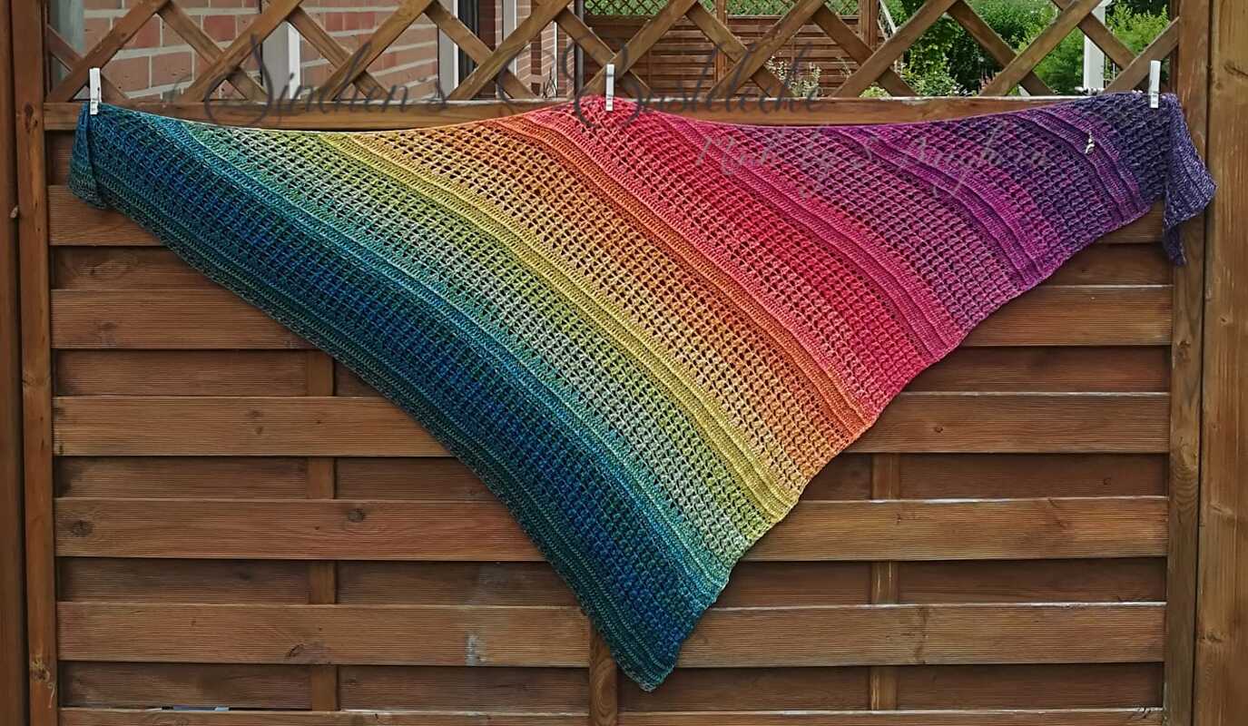 Triangle crochet shawl in rainbow gradient with openwork and textured stitch bands, clipped to a wooden fence.
