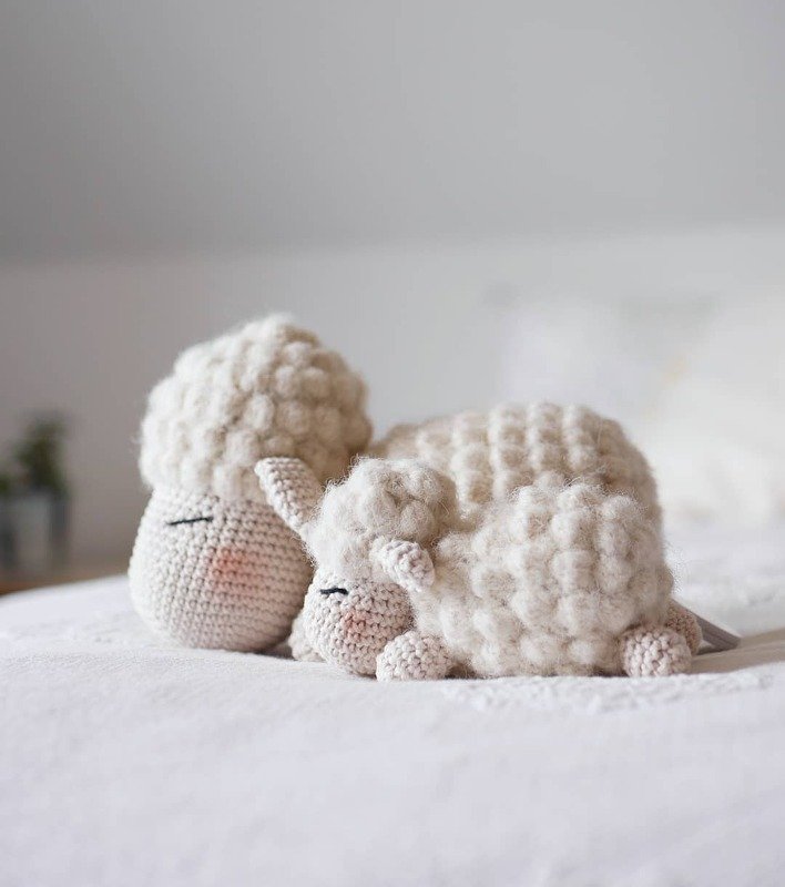 Two cream-colored crocheted sheep, one large and one small, sleeping on a white blanket.