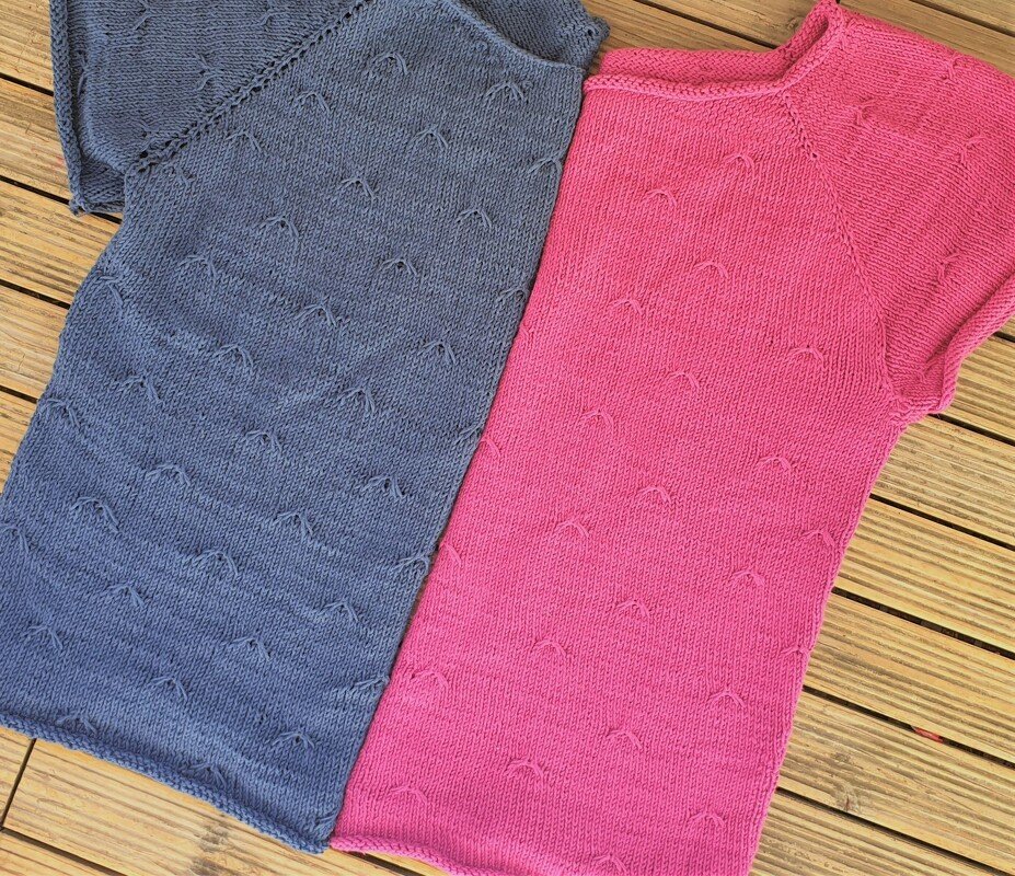 Close flatlay of two short raglan knit tunics showing crew neckline, raglan seam lines, and repeating arch eyelet stitch
