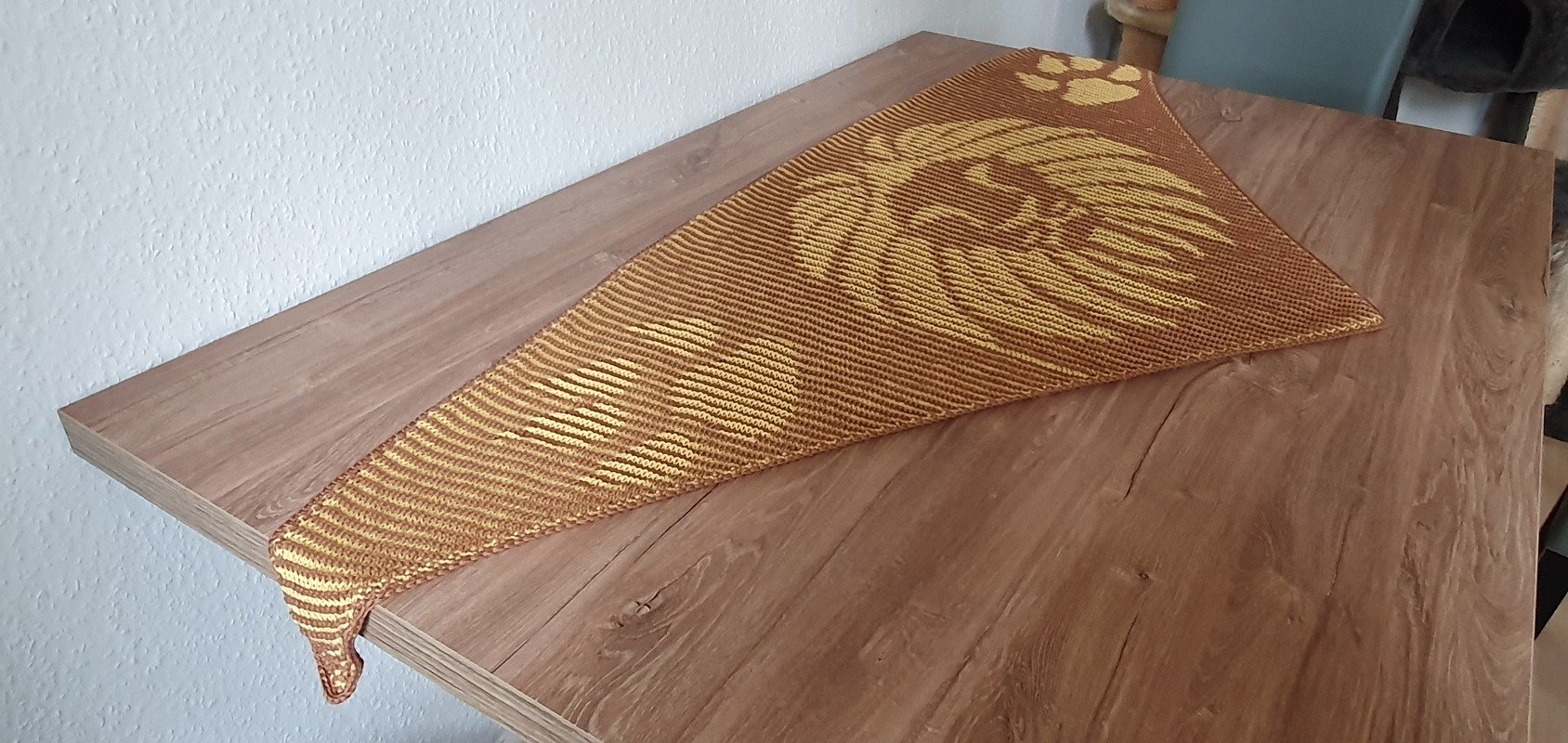 Triangular knitted shawl in brown and gold with a lion head motif and a paw print, displayed on a wooden table.