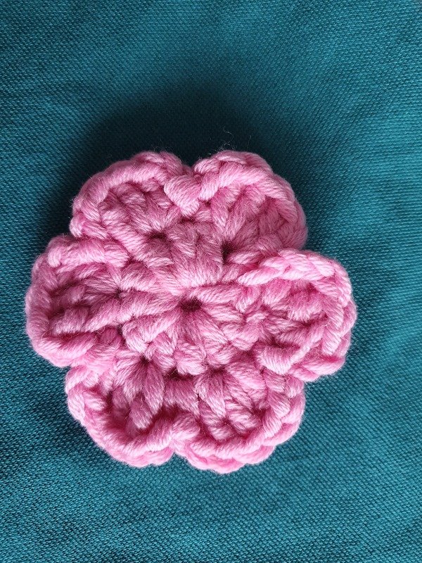 Nahaufnahme einer rosa gehäkelten Blüte mit sichtbaren Maschen auf petrolfarbenem Stoff