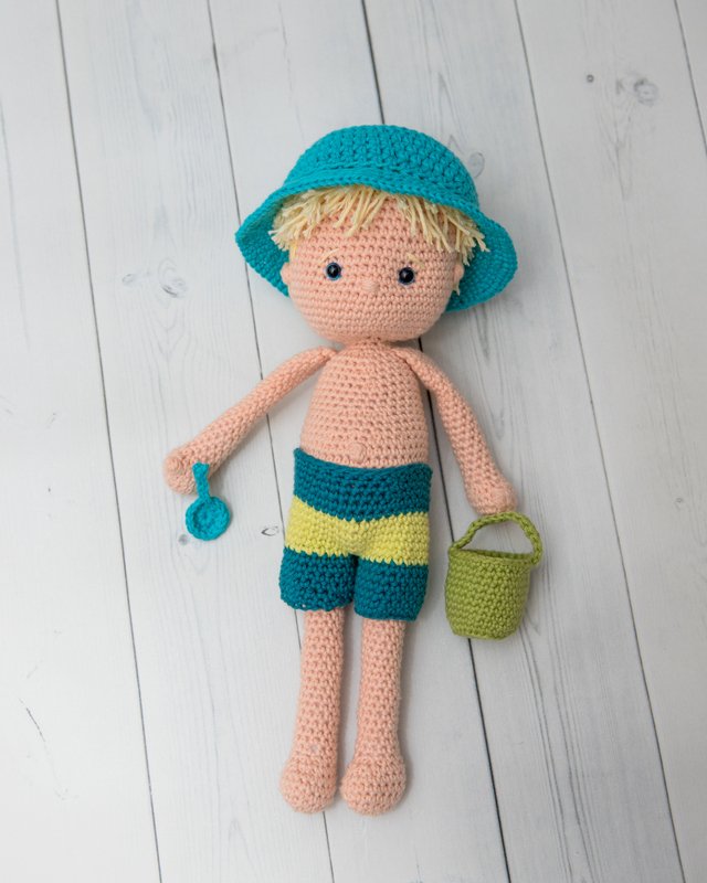 Gehäkelte Amigurumi-Puppe mit türkisem Hut, gestreiften Shorts, Eimer und Schaufel auf hellem Holzuntergrund